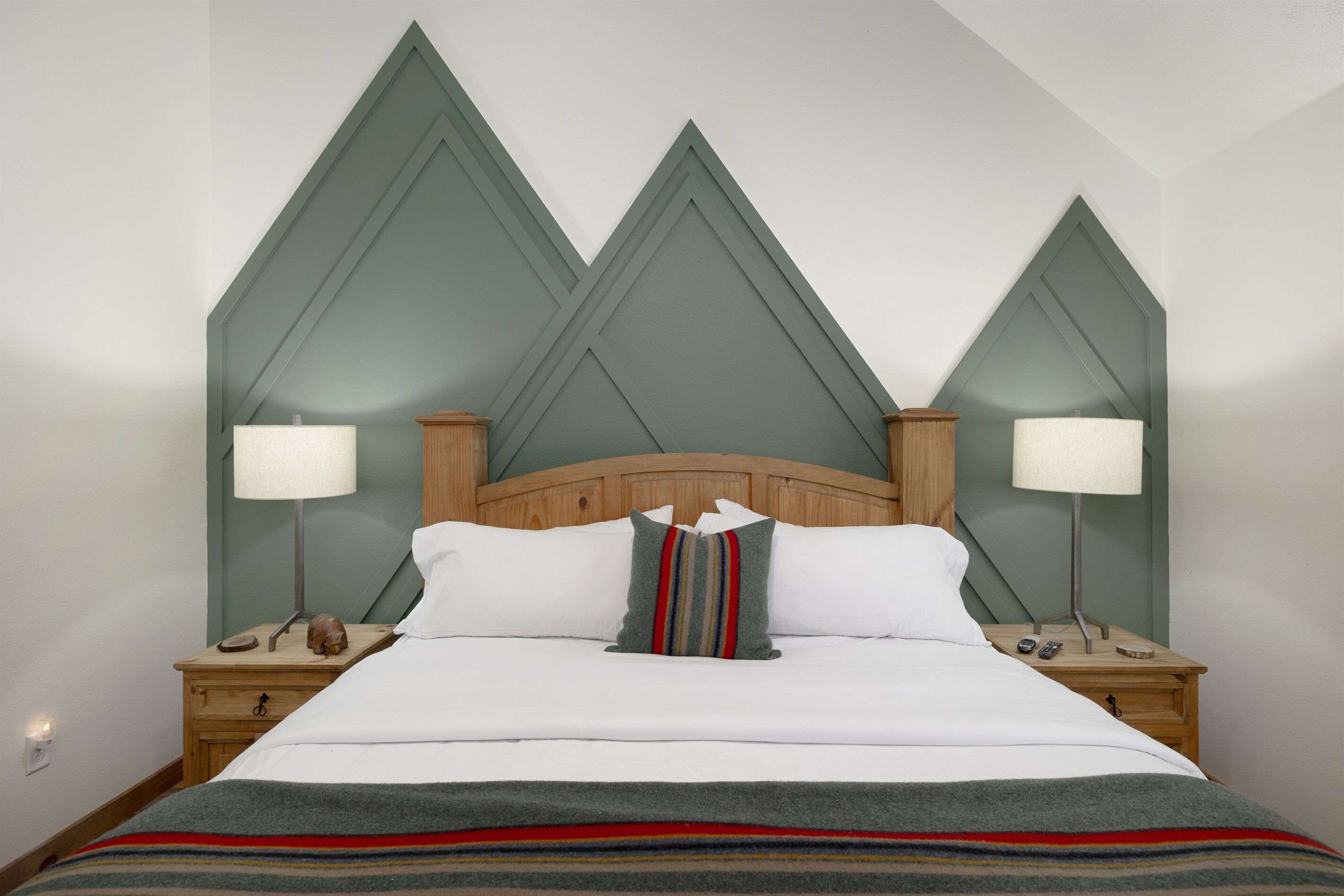 A neatly made bed with white linens, a striped throw blanket, and a pillow with a multicolored stripe. There are two bedside tables with lamps and a wooden headboard.
