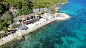 An aerial view of a coastal hotel with a pier extending into clear blue waters, surrounded by lush greenery and rocky cliffs.