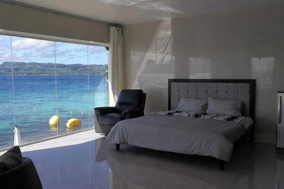 A bedroom with a large window, a bed, a chair, and a view of the sea.