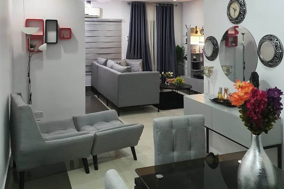 A modern living room with a grey sofa, a dining table with chairs, and a vase with flowers. There is a small table with a lamp and a wall-mounted air conditioner.