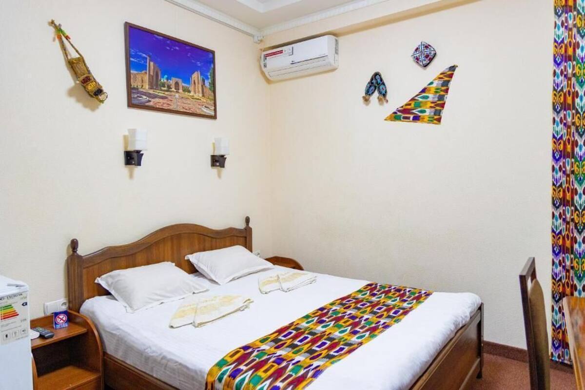 A hotel room with a wooden bed, white linens, a colorful blanket, a cityscape painting, an air conditioner, and decorative wall hangings.