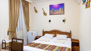 A hotel room with a bed, a desk, a chair, a nightstand, a wall-mounted air conditioner, and a painting of a cityscape. - Shakh Sultan Hotel (Tashkent)