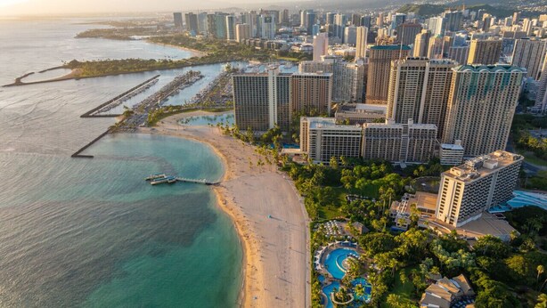 Vista aérea de un hotel frente al mar, con playa, piscina y edificios altos al fondo.