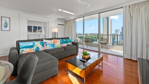 Una sala de estar moderna con un sofá gris, cojines coloridos, una mesa de centro de vidrio y vistas de la ciudad a través de ventanas amplias.