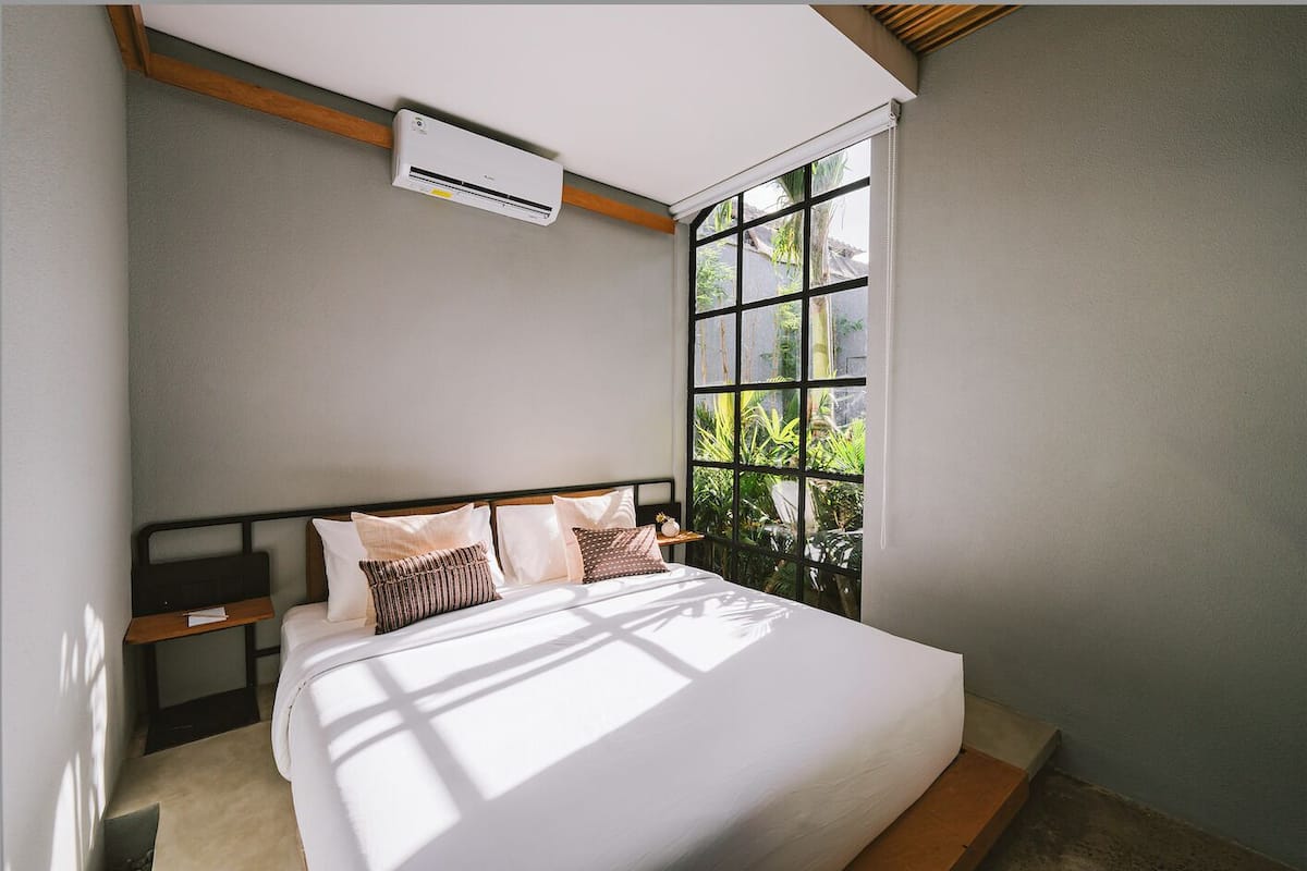 A bedroom with a large bed, a window with a view of greenery, and an air conditioning unit on the wall.