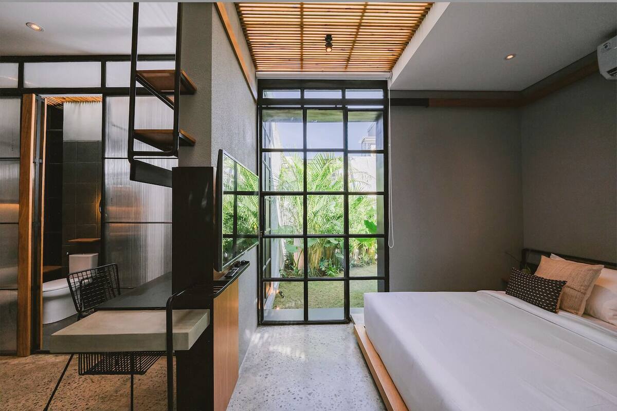 A modern bedroom with a large bed, a desk, and a view of greenery through floor-to-ceiling windows.