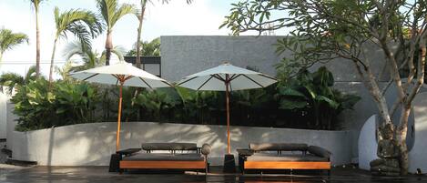 A poolside area with two lounge chairs, umbrellas, and a reflection of palm trees in the water.