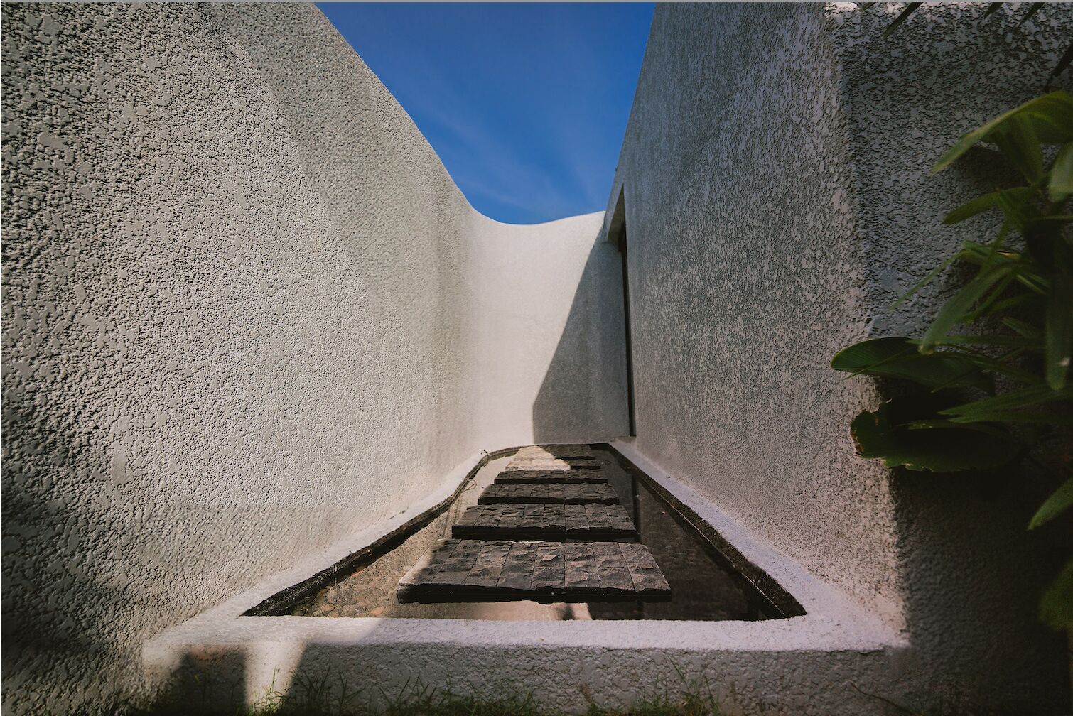A narrow alleyway with a stone pathway leading to a door, surrounded by textured walls and a clear blue sky above.