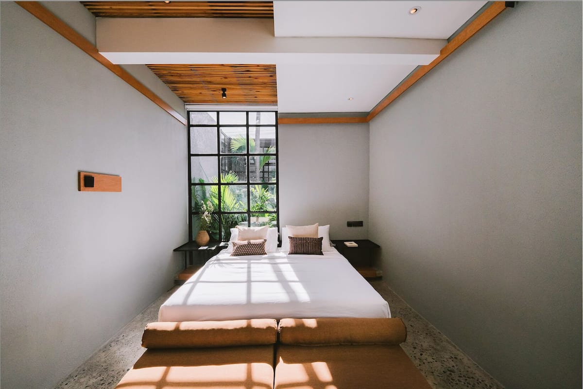 A bedroom with a large window, a bed, a nightstand, and a wooden ceiling beam.