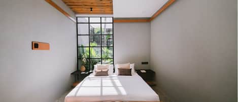A bedroom with a large window, a bed, a nightstand, and a wooden ceiling beam.