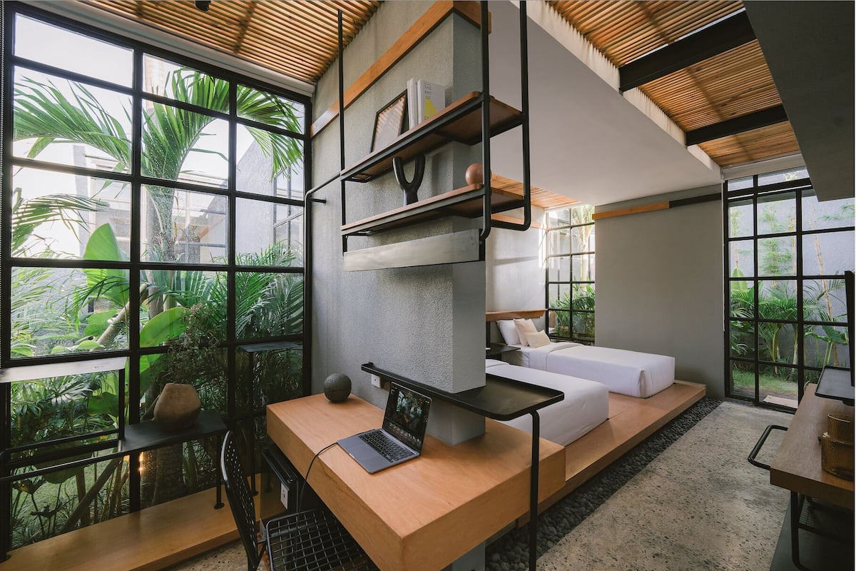 A modern interior with a wooden desk, a white sofa, and a bookshelf. Large windows provide views of greenery outside.