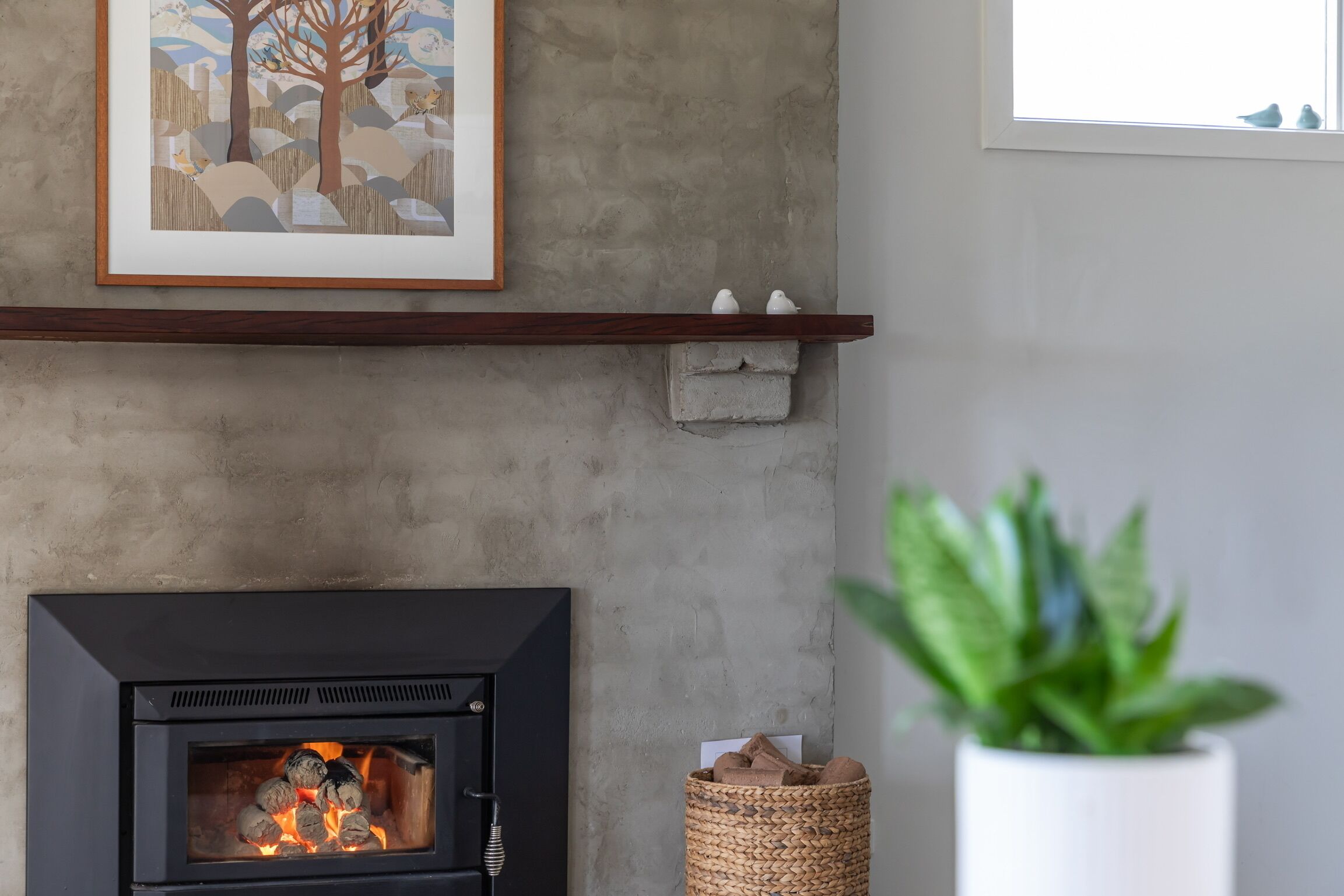 A fireplace with a framed picture above it, a shelf with decorative items, and a potted plant to the right.