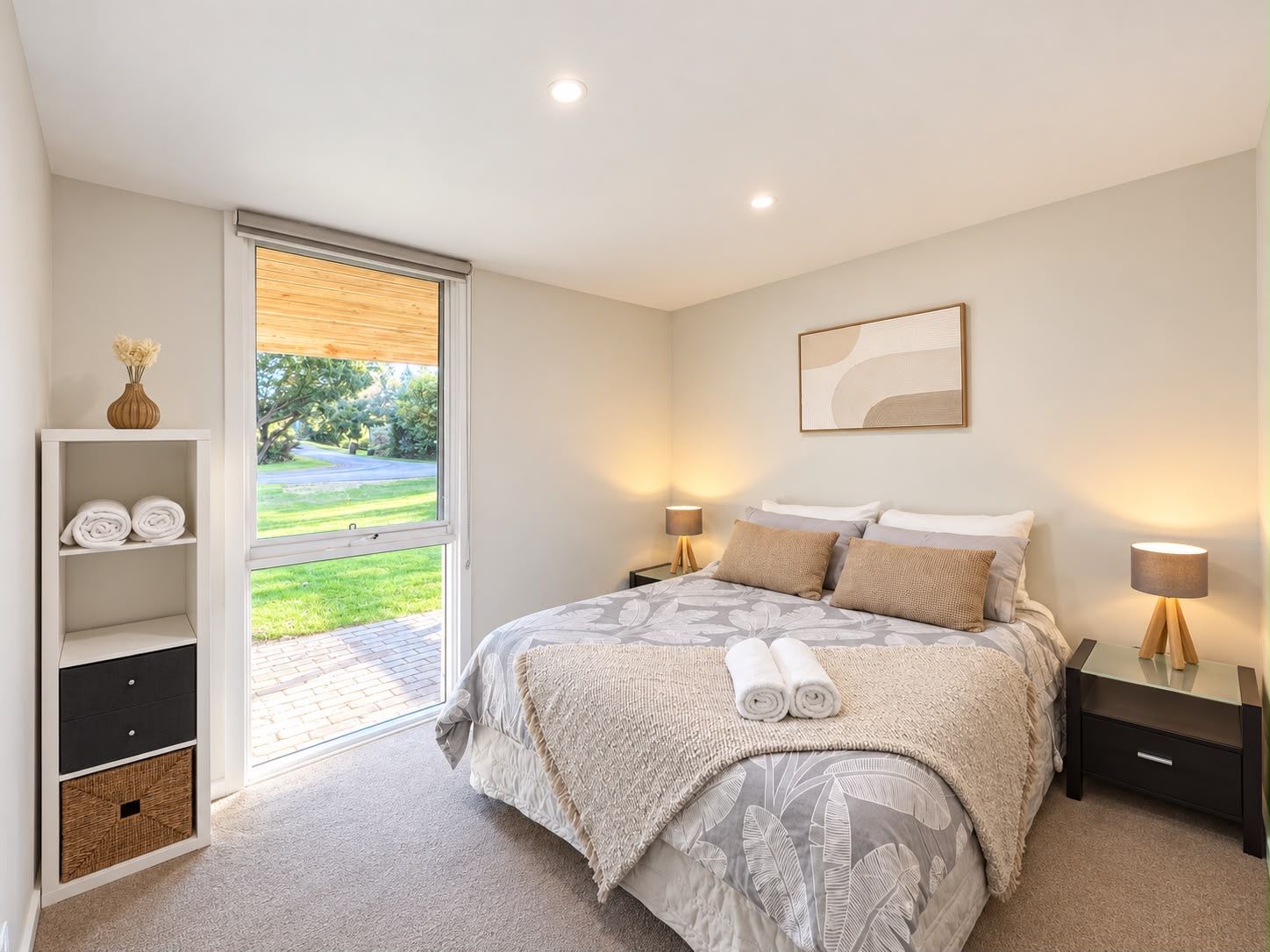A bedroom with a bed, bedside tables, a nightstand, a shelf with towels, and a view through a glass door.