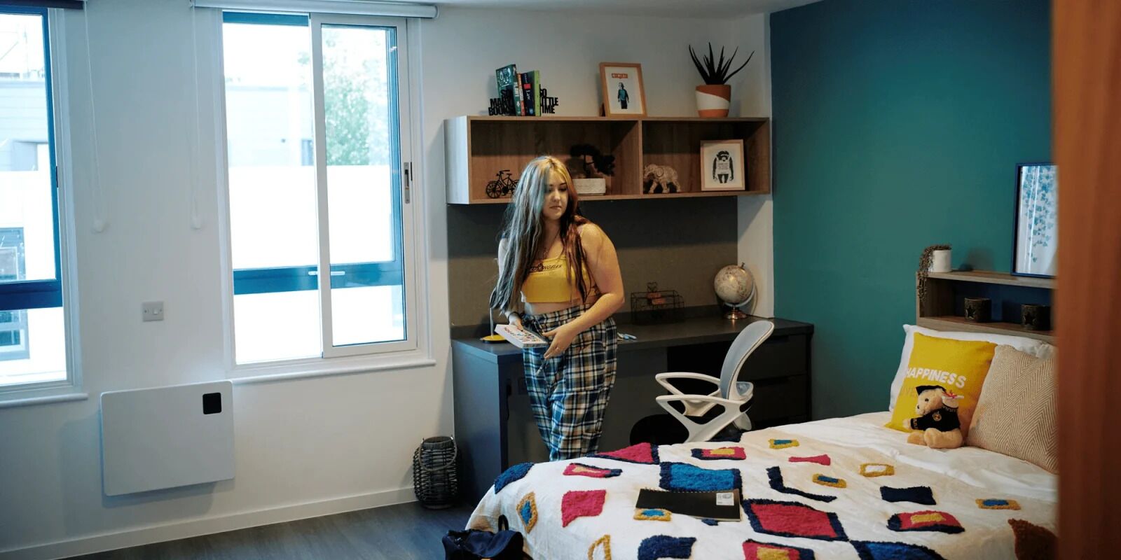 A room with a bed, a desk, a chair, a window, and a shelf with various items.