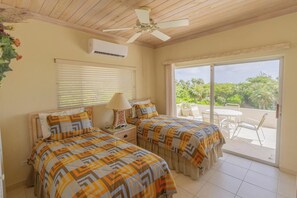 Un dormitorio con dos camas, ventilador de techo, aire acondicionado, ventana con persianas y un balcón con sillas y una mesa.
