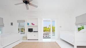 A bright, white room with a ceiling fan, a TV on a white cabinet, and a view of a pool and palm trees through an open door. - Caribbean Cliffside: Private Waterfront Retreat (North Palmetto Point)