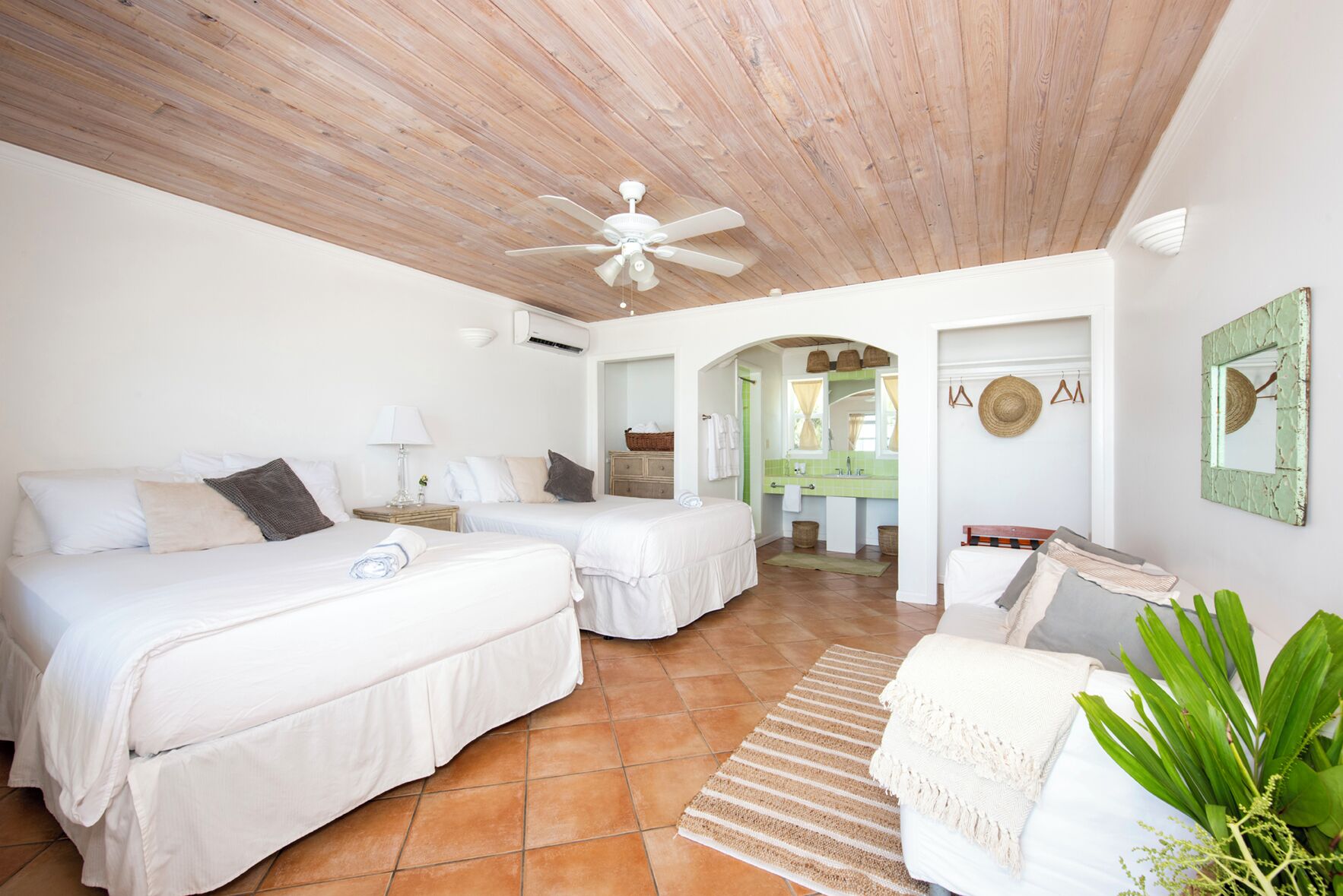 A hotel room with two beds, a ceiling fan, a bathroom with a green tiled wall, and a plant.