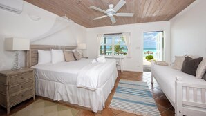 A bedroom with a large bed, a bedside table, a nightstand, a ceiling fan, a window with a view of the beach, and a balcony with a chair. - Windermere Wonder: Historic Home With Club Access (Savannah Sound)
