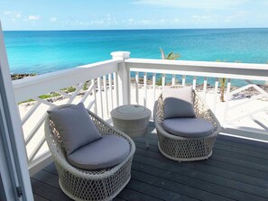A balcony with wicker chairs and a small round table, overlooking a beach with clear blue water and rocky shoreline. - Oceanfront Haven: Elegant Estate With Private Deck (James Cistern)