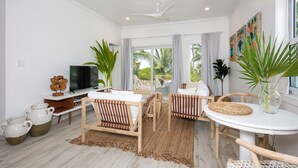 Ein modernes Wohnzimmer mit Fernseher, Sitzgelegenheiten und großen Fenstern mit Blick auf eine tropische Landschaft.