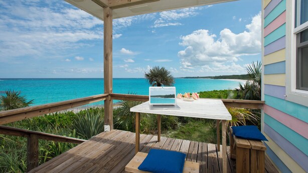Une terrasse en bois avec un ordinateur portable posé sur une table, donnant sur une mer turquoise et une plage.