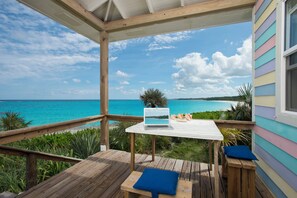 A wooden deck with a laptop on a table, overlooking a turquoise sea and a beach. - Cayo Loco Getaway: Private Beachfront Haven (Governor's Harbour)