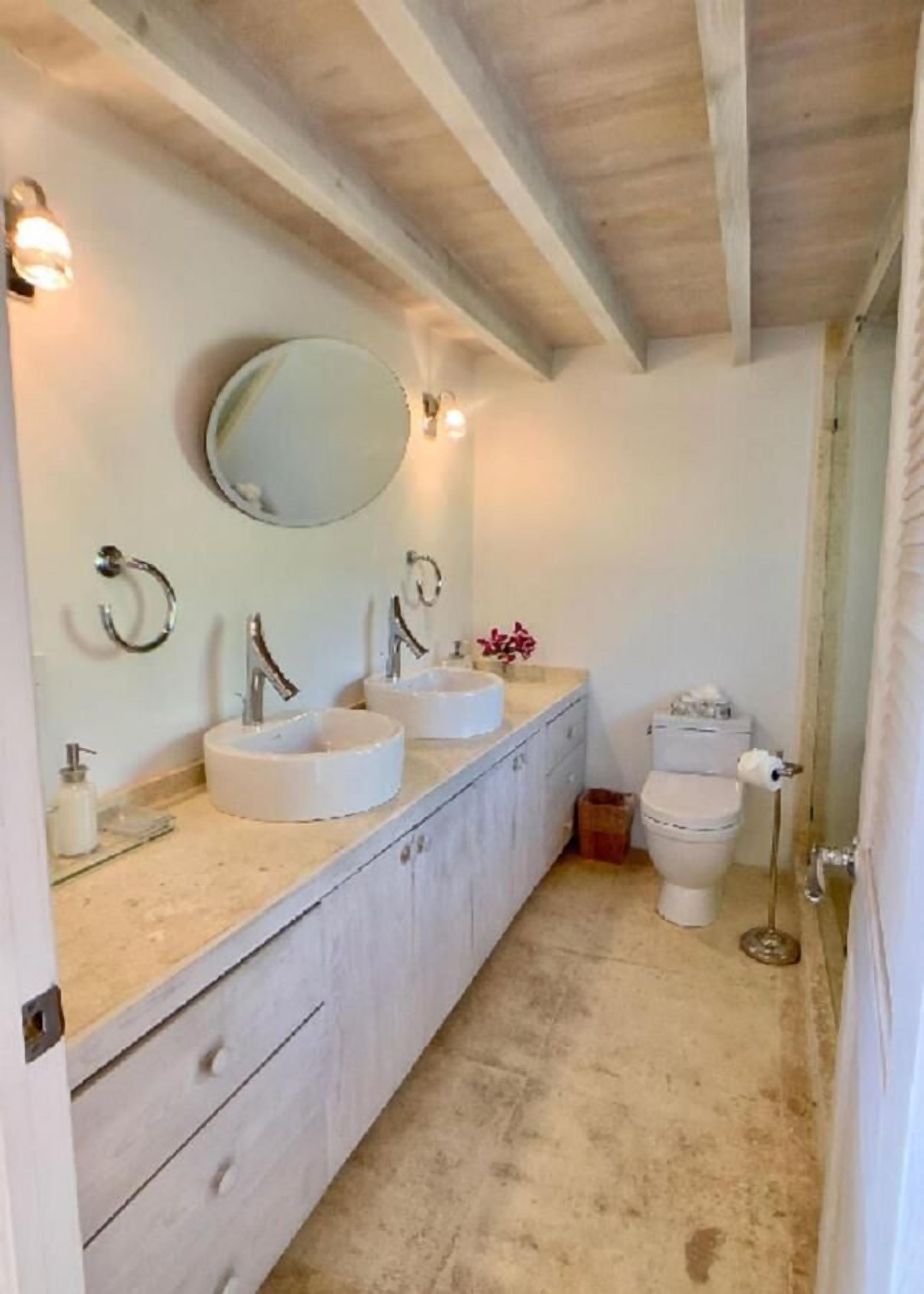 A bathroom with a long vanity, two sinks, a toilet, and a round mirror.