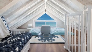 Ein gemütliches Schlafzimmer im Dachgeschoss mit schrägen Wänden, Meerblick, einem Bett mit gemusterten Textilien, einem grauen Pouf und einem weißen Babybett.