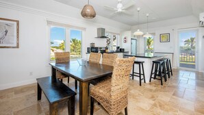A spacious dining area with a dark wooden table and woven chairs, a kitchen with modern appliances, and large windows offering views of palm trees. - Island Hideout: Quaint Cottage, Moments From The Beach (James Cistern)