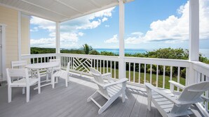 A covered outdoor area with white furniture, including chairs and a table, overlooking a scenic view of the ocean and greenery. - Island Hideout: Quaint Cottage, Moments From The Beach (James Cistern)