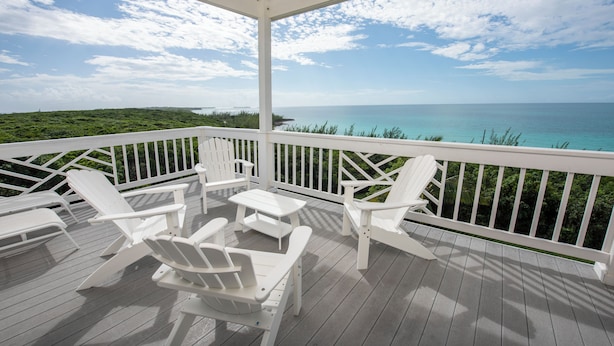 Une terrasse extérieure couverte, dotée de chaises Adirondack blanches et d’une table, offrant une vue sur un océan turquoise et un paysage verdoyant.