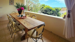 Comedor con mesa de madera y sillas de mimbre, ventilador de techo y vistas a la vegetación y al mar a través de una ventana.