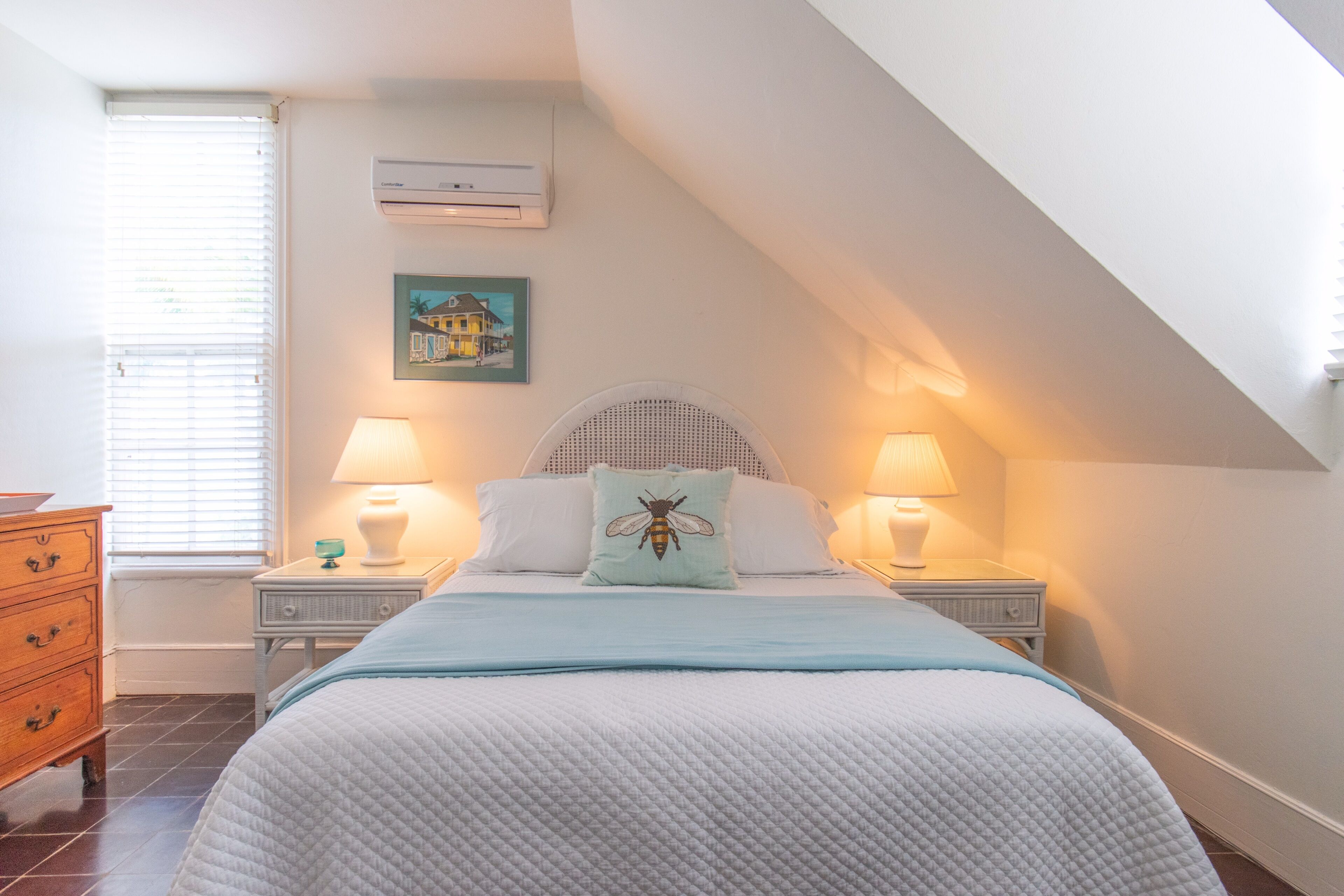 A bedroom with a bed, two bedside tables with lamps, a dresser, a window with blinds, an air conditioner, and a wall-mounted picture.