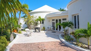 A white building with a curved roof, a patio area with white chairs, and palm trees. - Exquisite Retreat: Contemporary Estate On Pink Sand Beach (Double Bay)