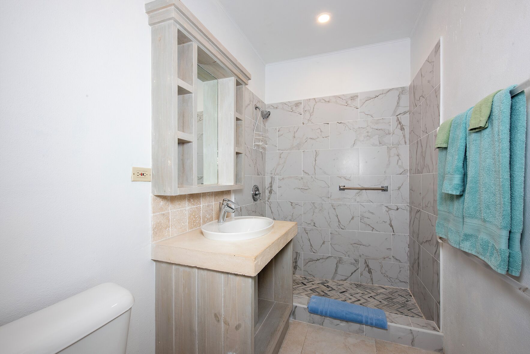 Un baño con pared de ducha de mármol, lavamanos con tocador de madera y un perchero con toallas.