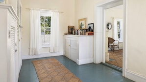 A hallway with a patterned rug, a white door with shutters, a white cabinet with books, and a mirror reflecting a chair and window. - Captain'S Cove: Historic House Near Town And Beach (Governor's Harbour)