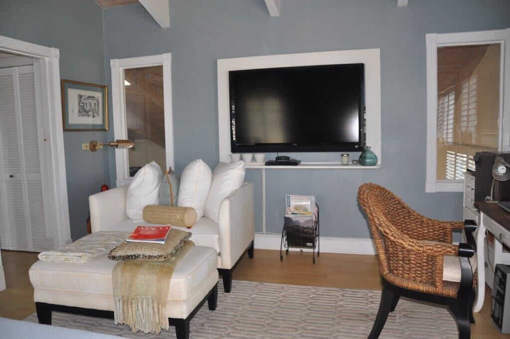 Una sala de estar con un sofá blanco, un sillón de mimbre, un televisor de pantalla plana montado en la pared y una mesita auxiliar con una revista.