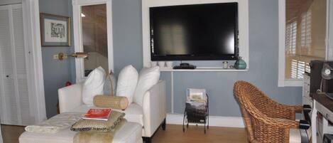 Una sala de estar con un sofá blanco, un sillón de mimbre, un televisor de pantalla plana montado en la pared y una mesita auxiliar con una revista.