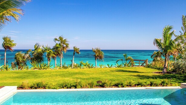 A pool with clear blue water, surrounded by a well-maintained lawn and palm trees, overlooking a calm sea. - Wild Orchid Hideaway: Lux Beachfront Sanctuary (Governor's Harbour)