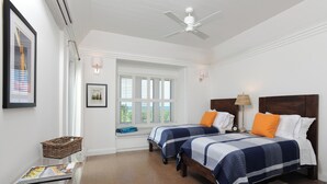 A bedroom with two beds, a ceiling fan, a window with blinds, a lamp, and a framed picture on the wall. - Coastal Haven: Charming Beachfront Cottage With Pool (Governor's Harbour)