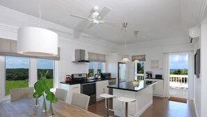 A modern kitchen with white cabinetry, a central island, and a dining area with a wooden table and chairs. - Coastal Haven: Charming Beachfront Cottage With Pool (Governor's Harbour)