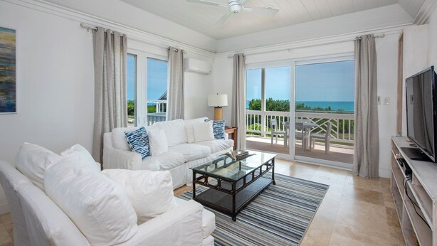 A spacious living room with a white sofa, a glass coffee table, and a TV on a wooden stand. The room has large windows with curtains, a ceiling fan, and a view of the ocean. - Oceanfront Bliss: Newly Built Luxury Home With Sunset Views (North Palmetto Point)