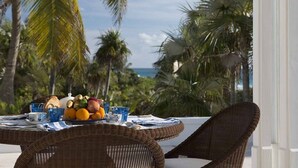 Ein Korbtisch mit Frühstücksdeko, darunter Obst, Brot und Tassen, mit Blick auf einen tropischen Strand mit Palmen und dem Meer.