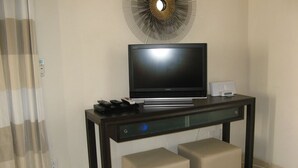 A modern hotel room with a flat-screen TV on a dark wooden console table, two beige ottomans, and a decorative wall piece above the TV. - Caribbean Retreat: Private Villa With Panoramic Ocean Views (Governor's Harbour)