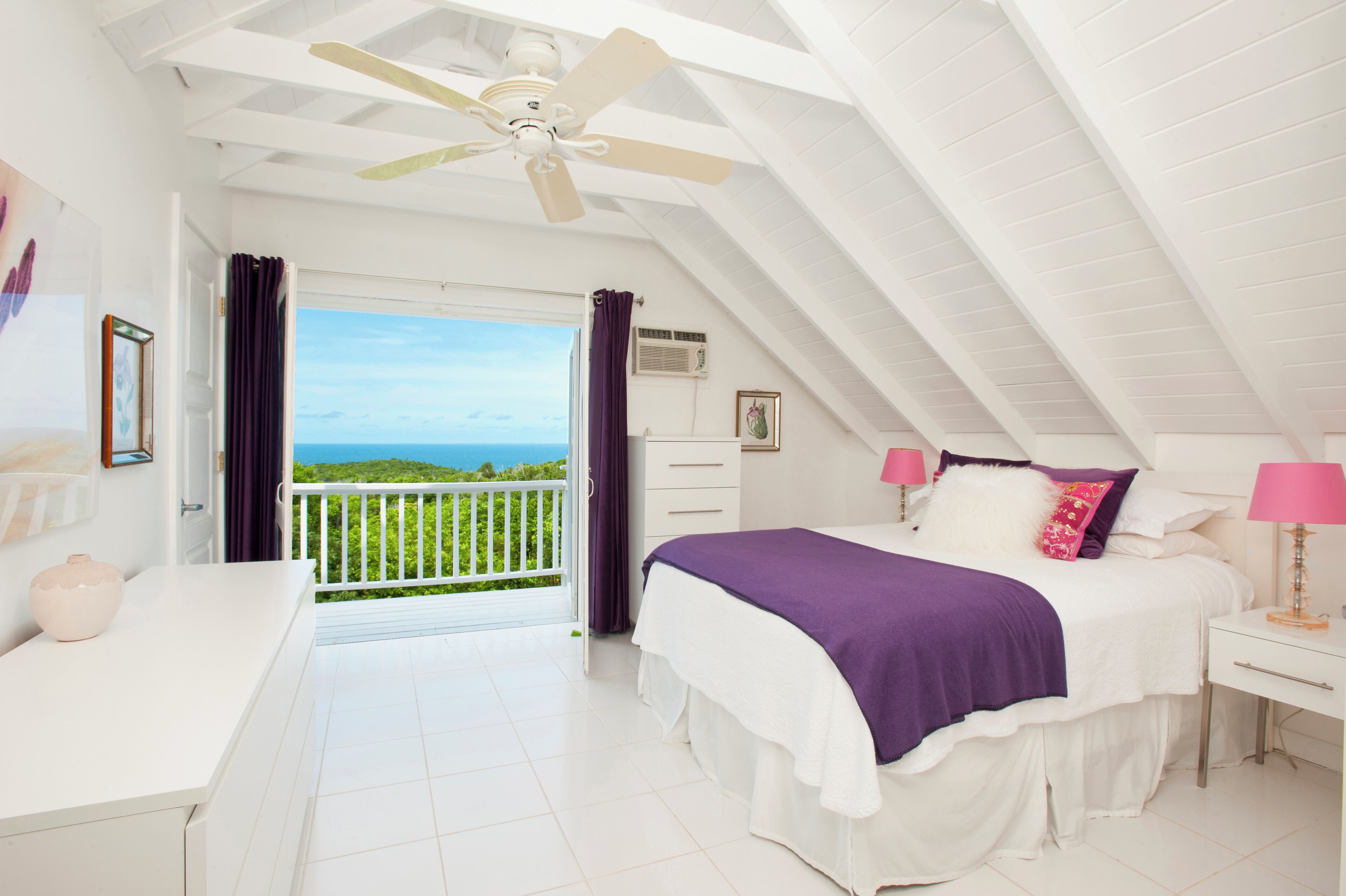 Une chambre à coucher avec un lit, une table de chevet, un ventilateur de plafond, une fenêtre avec des rideaux et une vue sur l’océan.