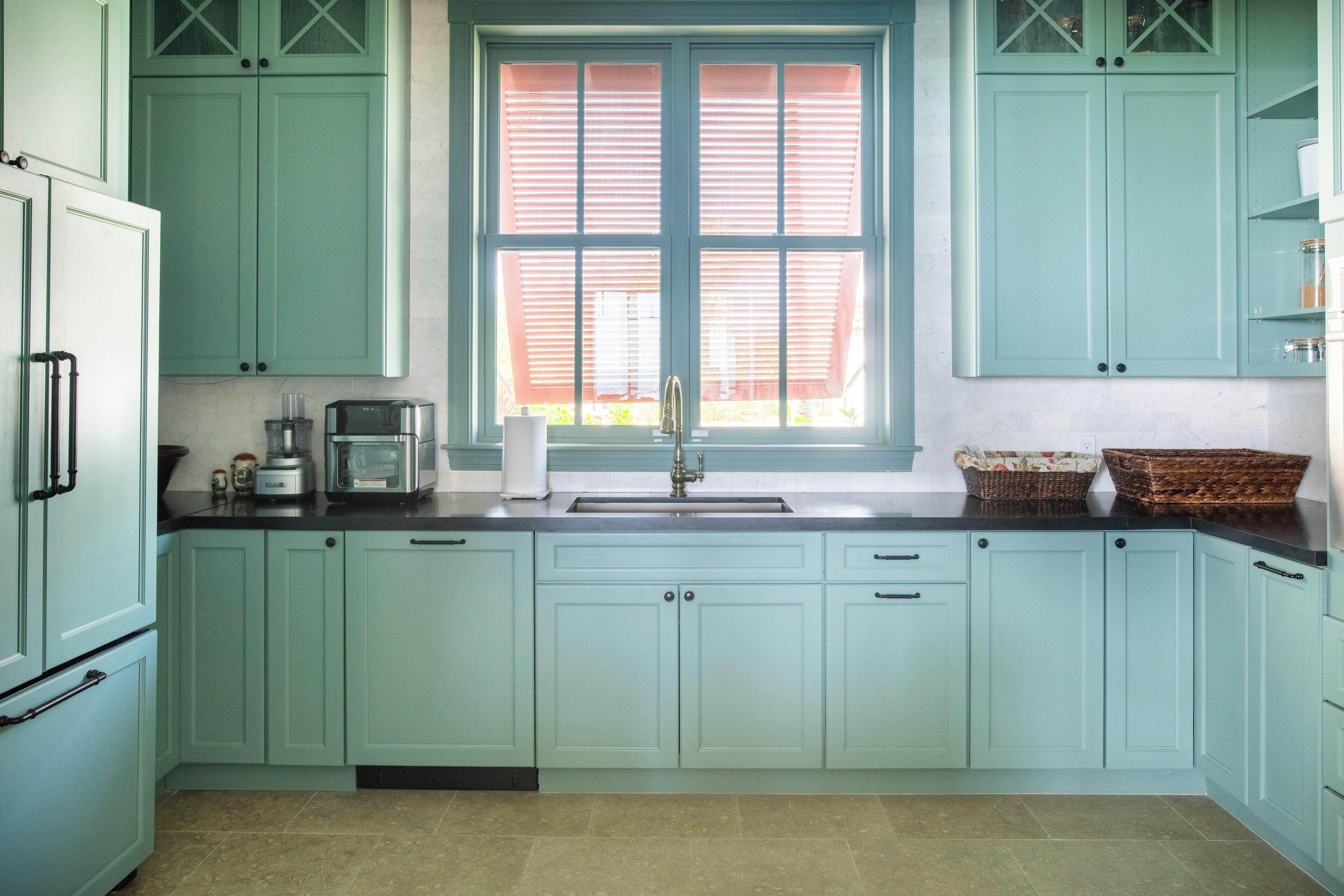 A kitchen with light green cabinets, a window with blinds, a sink, and a countertop with a coffee maker and toaster.