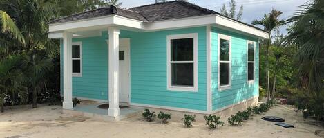 Una casa pequeña de un solo piso con fachada turquesa y detalles blancos, rodeada de vegetación tropical.