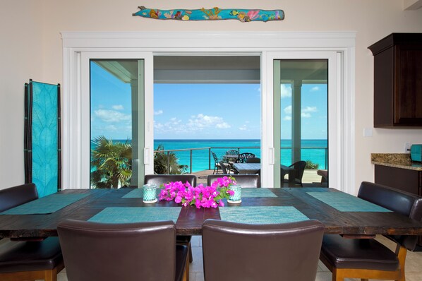 A dining area with a view of the ocean, a table with chairs, and a decorative hanging above. - Buttonwood Bliss: Lux Condo With Views (James Cistern)