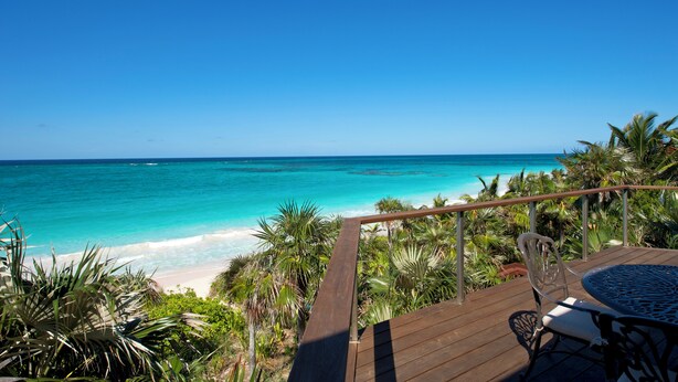 A wooden deck with a table and chairs overlooking a beach with turquoise water and palm trees. - Buttonwood Bliss: Lux Condo With Views (James Cistern)