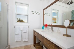 A bathroom with a white vanity, a large mirror, and a window with a view of palm trees. - Touch Of Class Cottage: Beachfront Oasis (Savannah Sound)